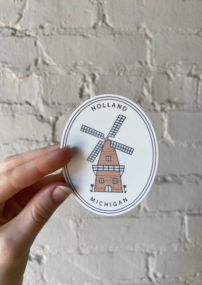 Hand holding an oval sticker in front of a white brick wall. Sticker displays a windmill with the words Holland Michigan on it