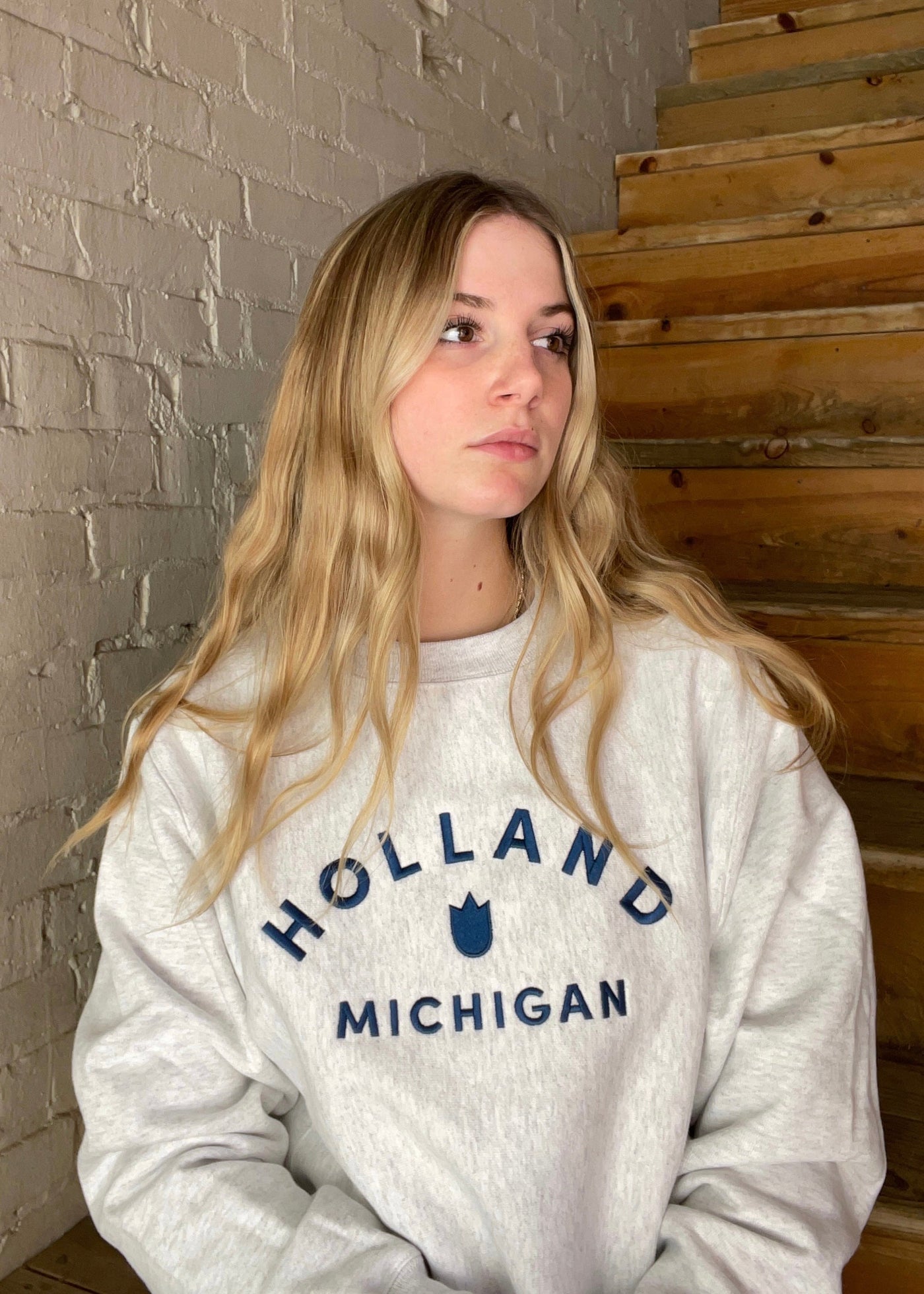 Blond female model wearing a heather grey crewneck sweatshirt with Holland Michigan and a tulip in navy embroidered on it.