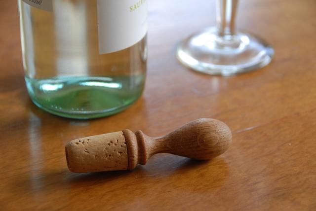 An oak wine stopper in an oval shape laying in front of a wine bottle and glass.